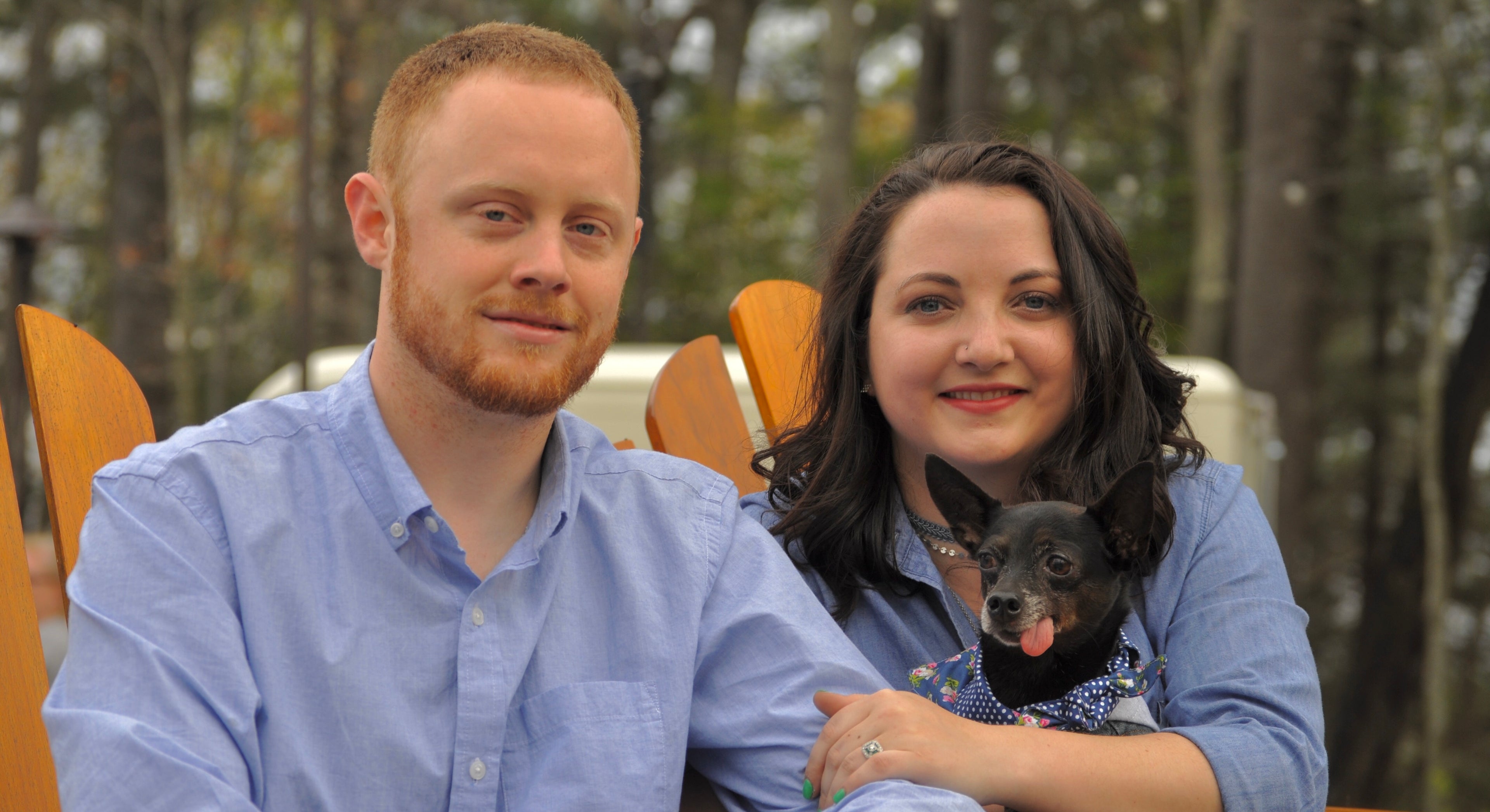 Man and woman sitting outdoors with a small dog, smiling at the camera.